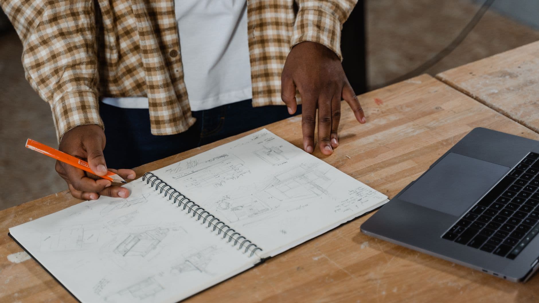 A sketchbook of hand drawn plans open on a workbench beside a laptop, with the maker's hands resting on the bench