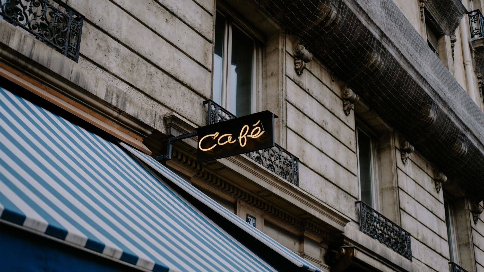 A small illuminated cafe sign mounted on a stone building above a striped awning