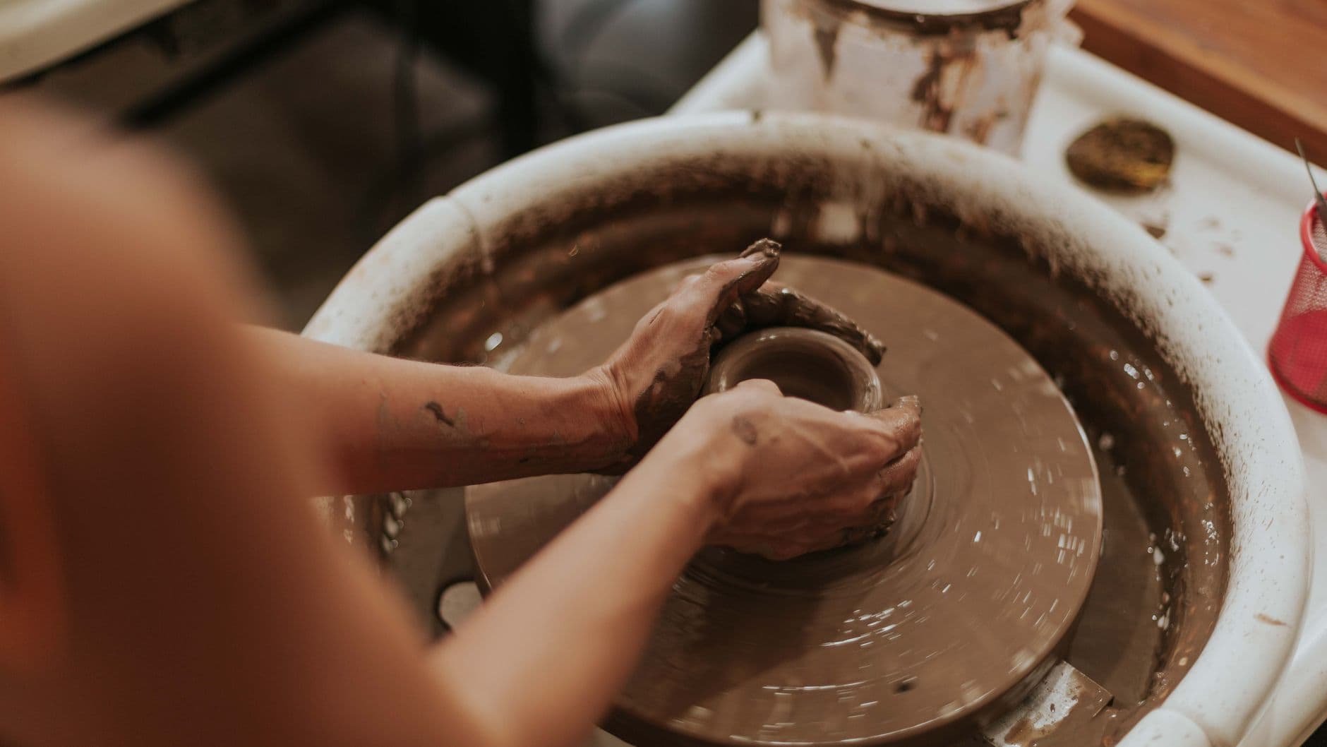 A potter's clay covered hands shaping the wall of a bowl on a spinning wheel